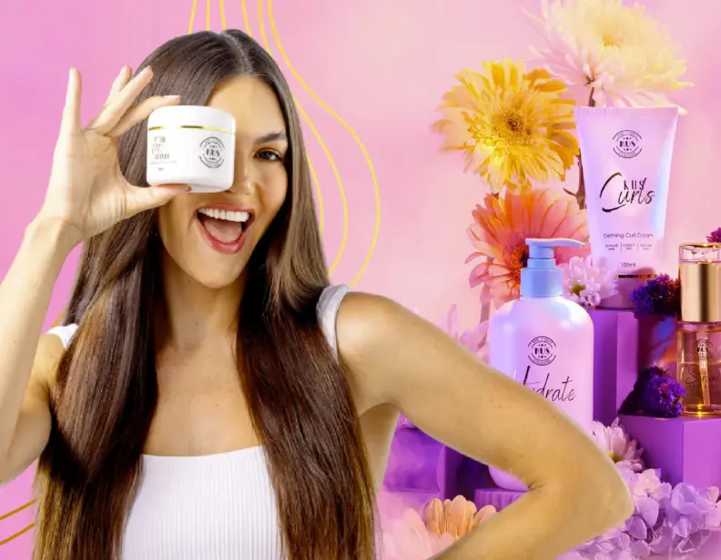 Smiling woman with long dark hair holding a white skincare jar near her eye, with skincare products and flowers in the background on a pink backdrop.